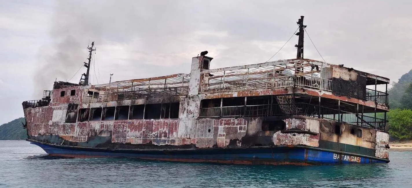 The burnt hull and upper decks of a passenger ferry, still smoking, in calm water near a green coast.