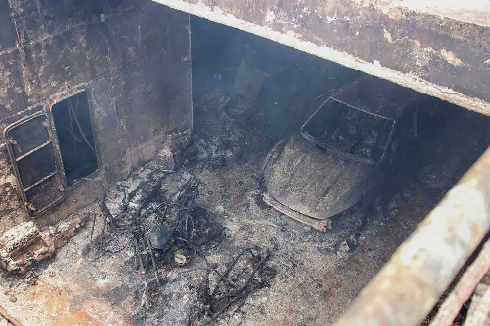 A burnt car and the burnt metal frames of several motorbikes in a dark, fire-damaged cargo space.