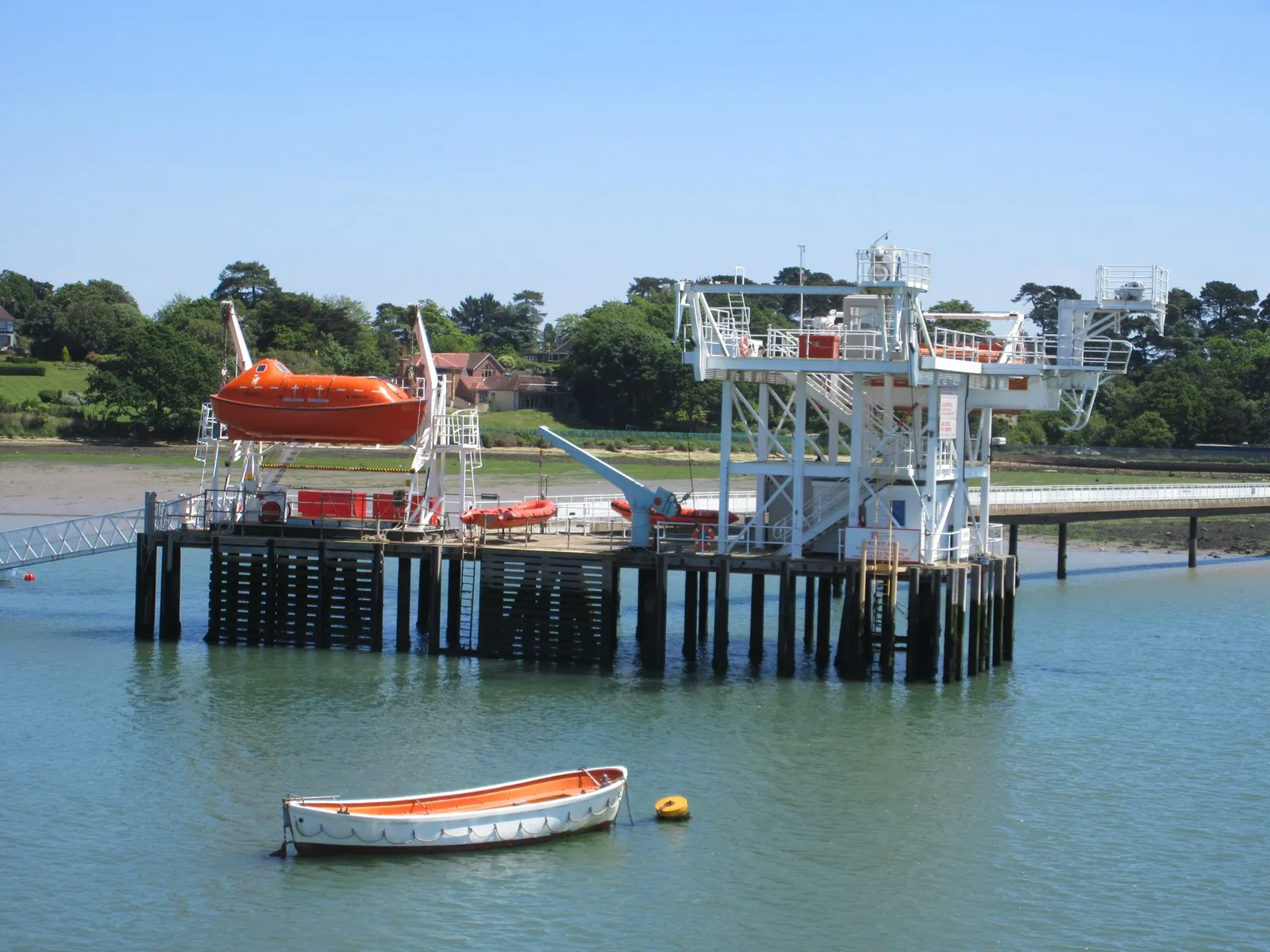 An orange enclosed lifeboat hangs from a white steel launching structure on a pier over calm water, with a small open boat moored in front.