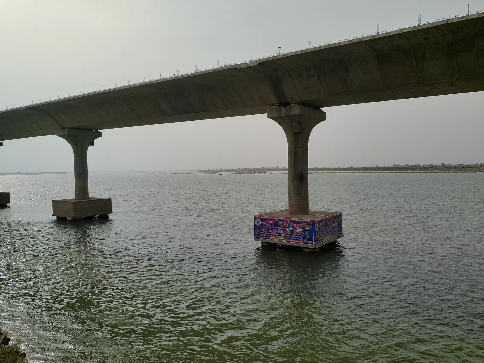A long concrete bridge on tall pillars crosses a wide, calm river under a grey sky.