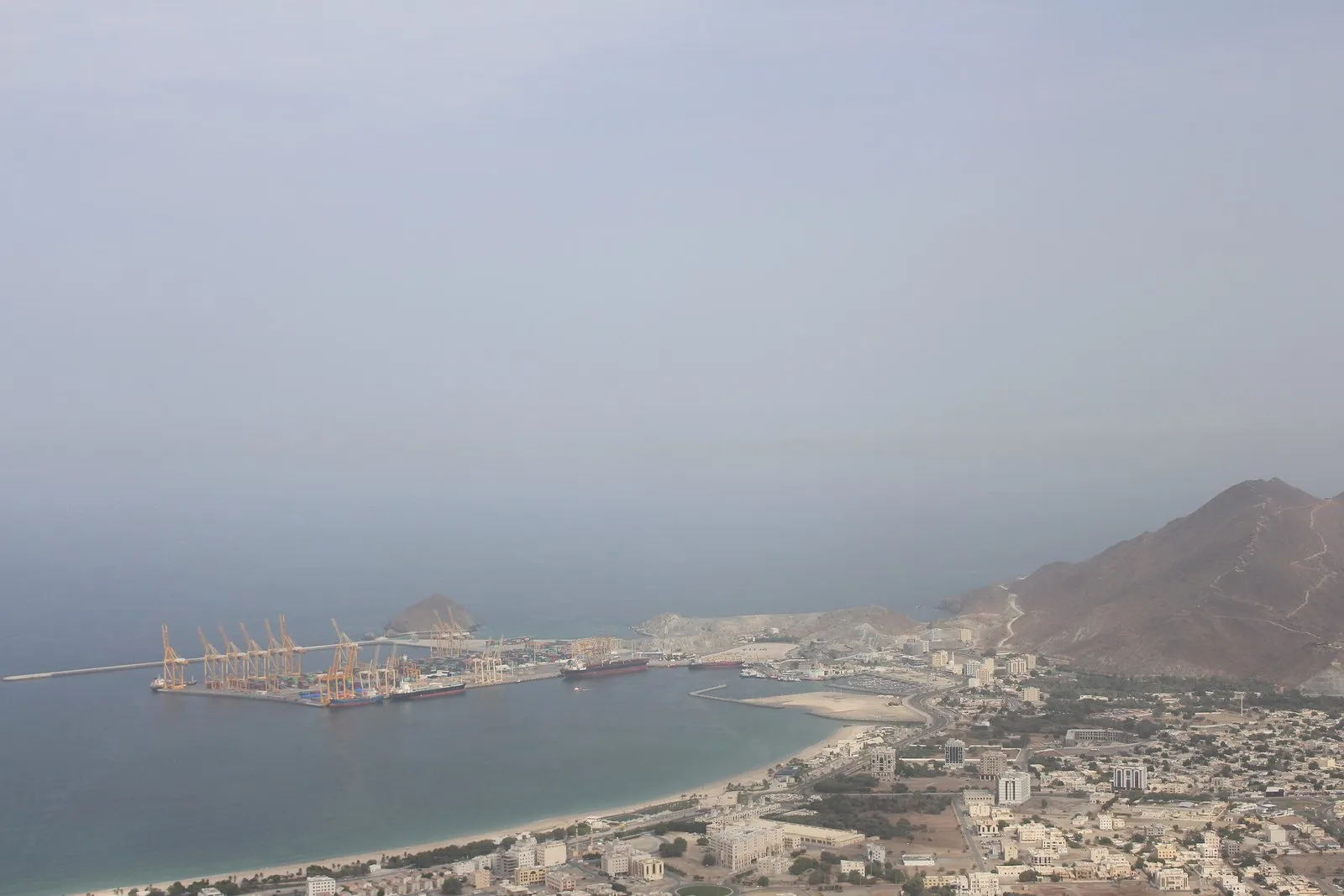 Hazy view over the town of Khor Fakkan, its bay and the container port, with mountains on the right.