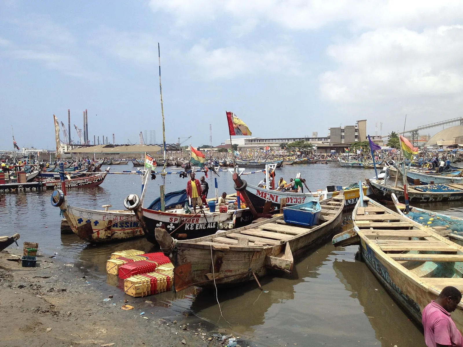 Many colourful wooden fishing canoes with flags in a harbour. Some fishermen stand on the boats and on the shore.