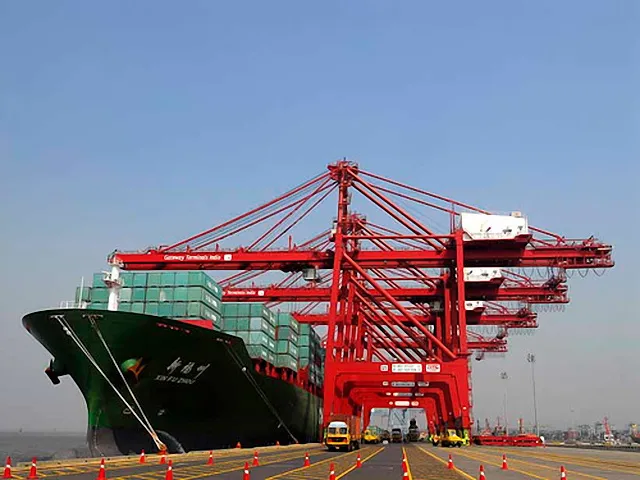 A large green-hulled container ship loaded with containers, moored under tall red cranes, with orange traffic cones on the quay.