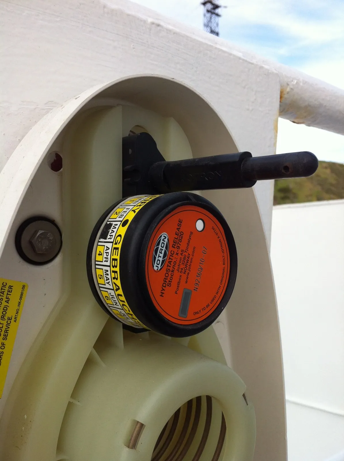 A round orange and black release unit with a date label fixed to a white plastic beacon bracket on a ship's railing.