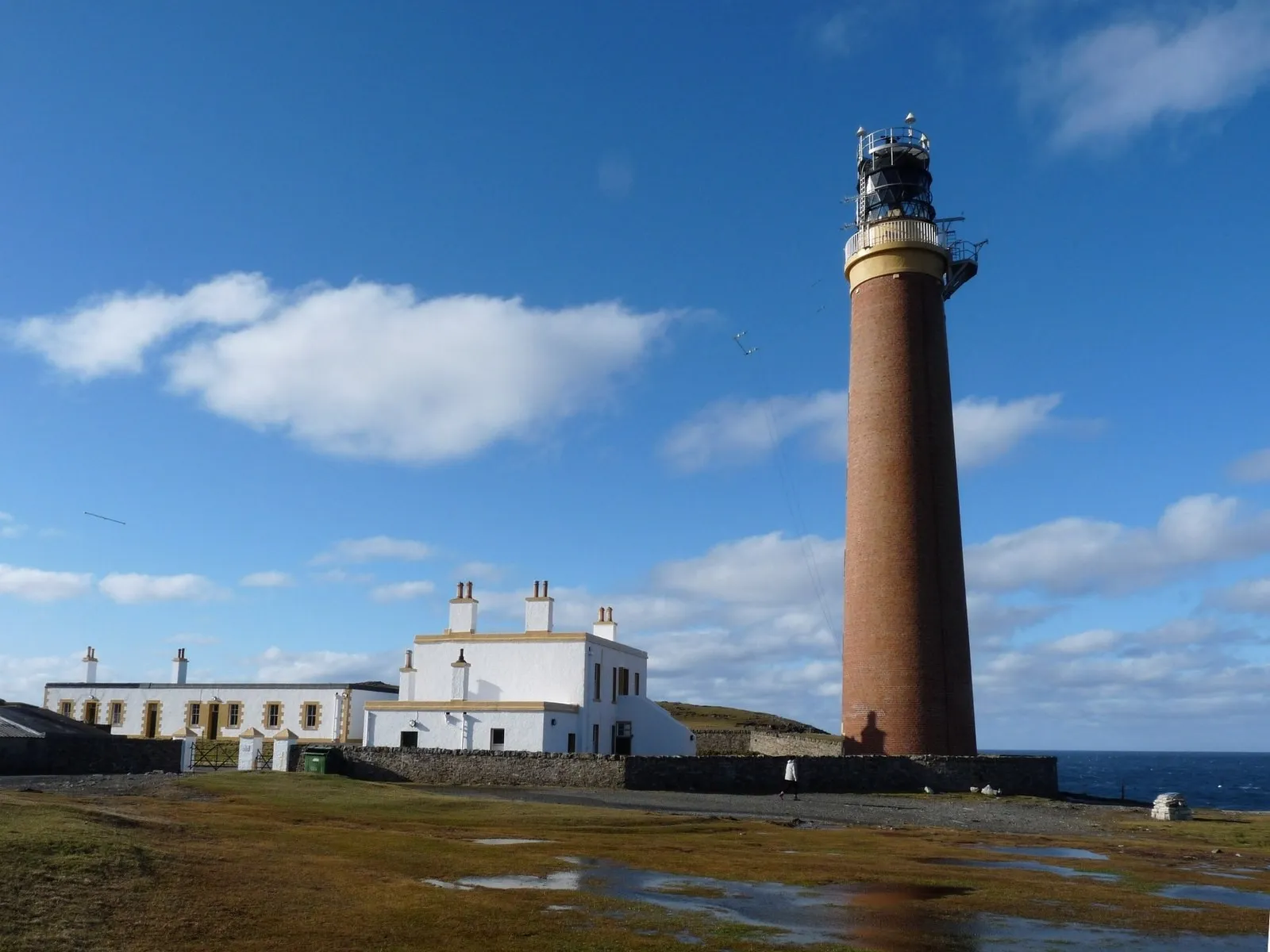 A tall red-brick lighthouse next to white buildings on flat grassland, with the sea behind it.