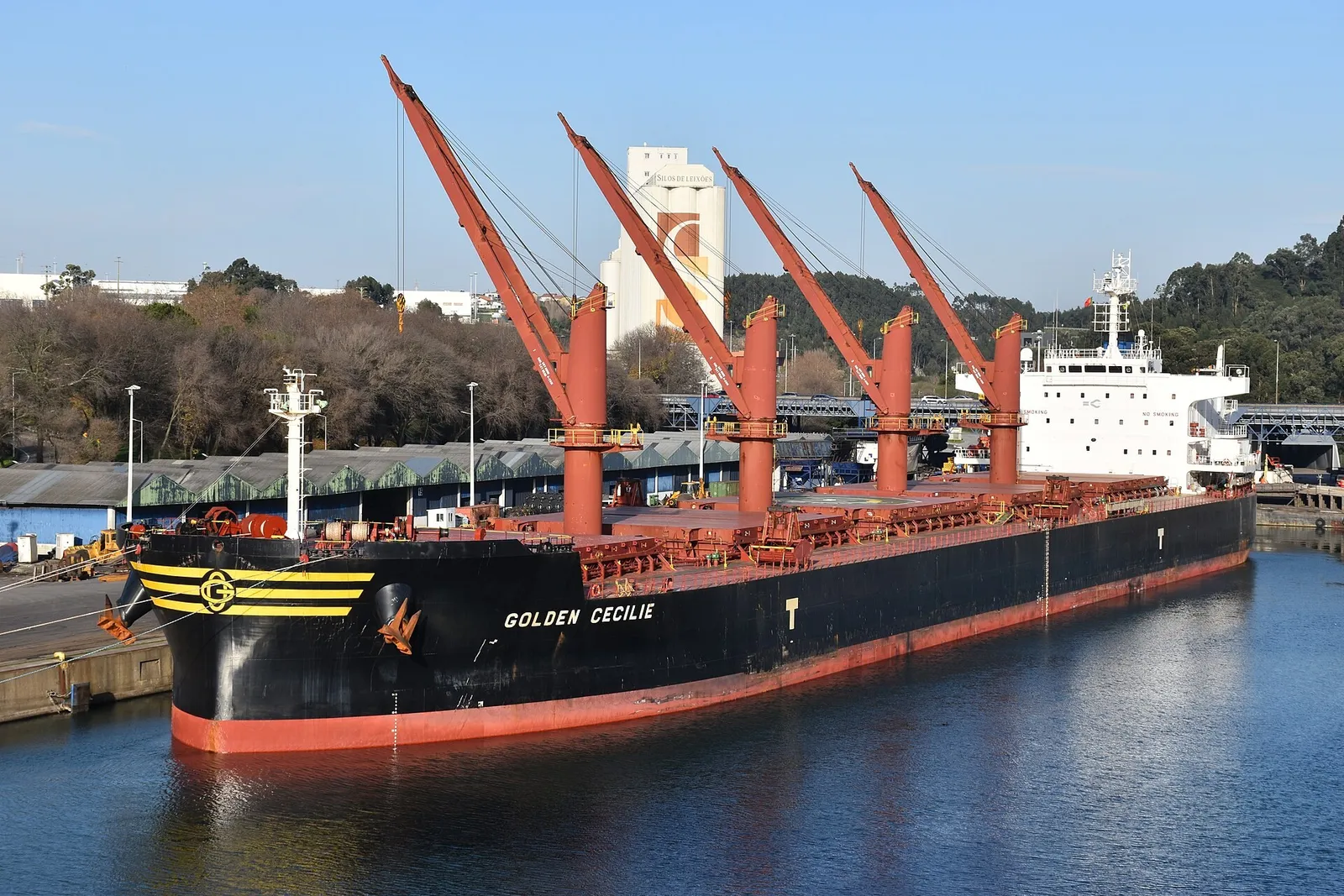 A black bulk carrier with four orange deck cranes raised, moored at a quay.