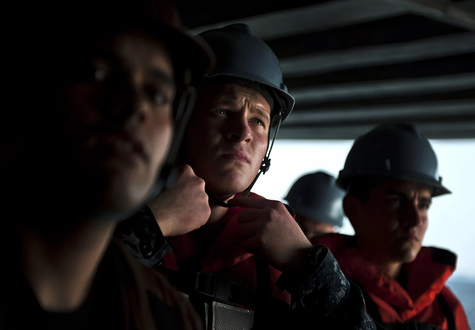 A sailor in a grey hard hat and red life jacket pulls the chinstrap tight under his chin, with other crew behind him.