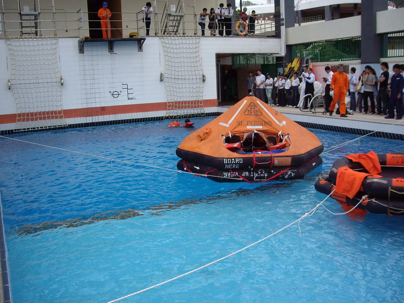 Two orange life rafts float in a blue indoor training pool. People in orange suits and other trainees stand at the side of the pool.