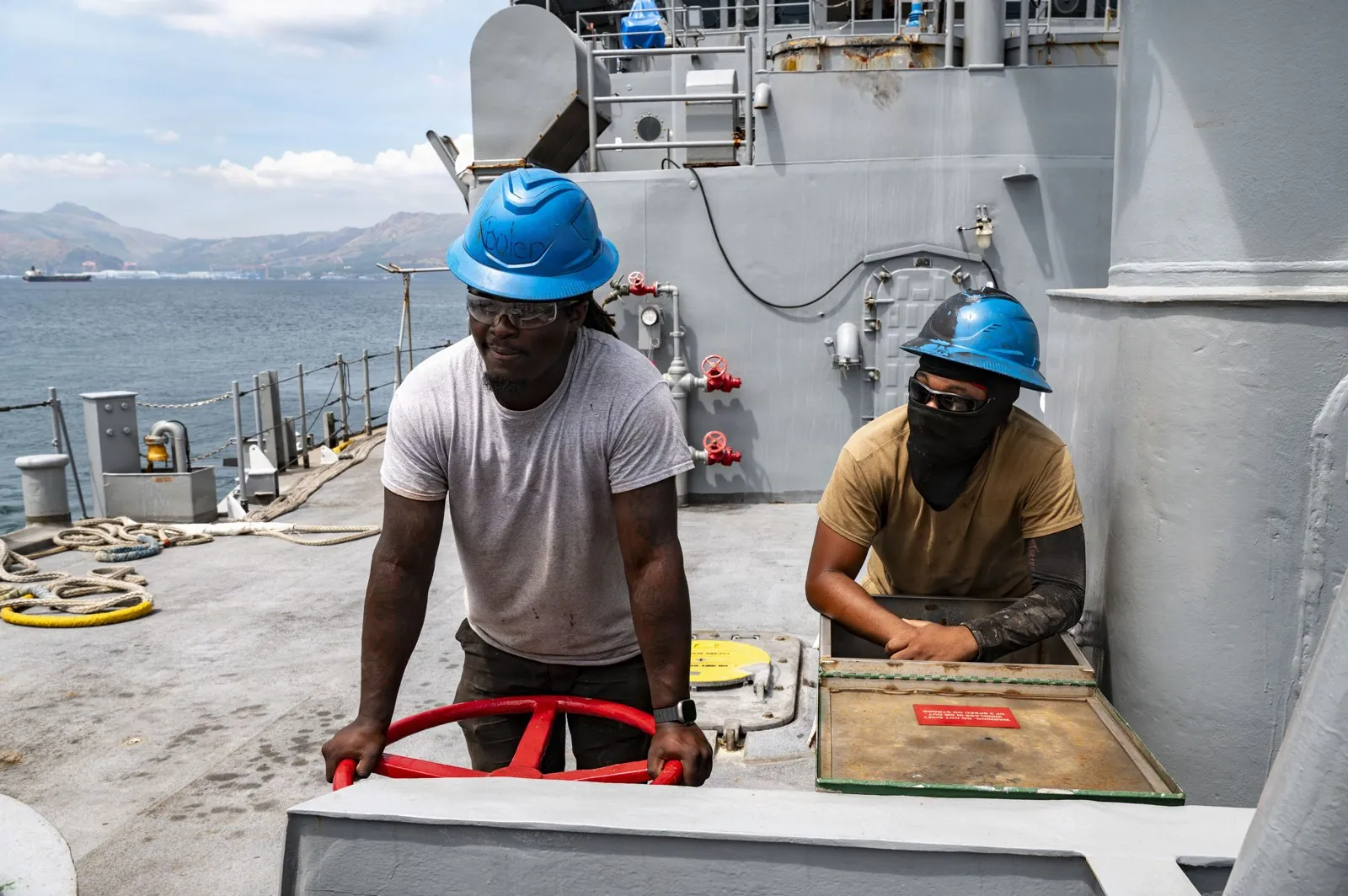Two crew members in blue hard hats work at a red wheel and an open deck box on the grey foredeck of a ship, with the sea and hills behind.