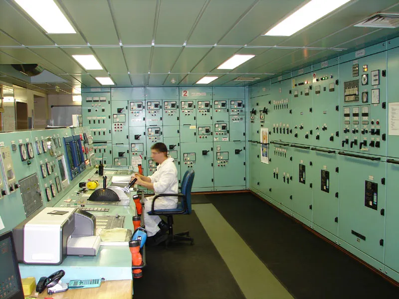 A crew member in white overalls sits at a control desk in a ship's engine control room with many green panels and gauges.