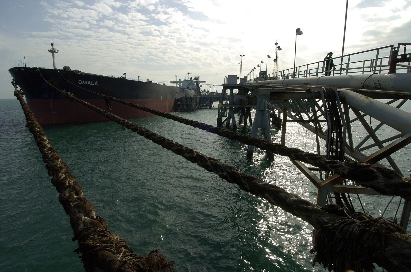 The dark hull of a large oil tanker lies next to a steel oil terminal, with thick mooring ropes in the front.