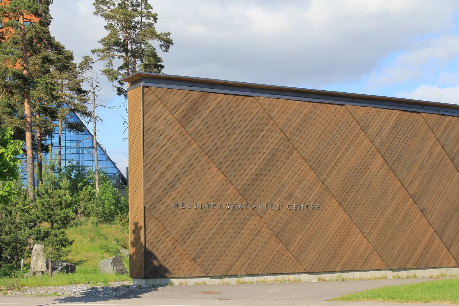 A wooden-clad building with the words Helsinki Seafarers' Centre on its wall, with pine trees and a glass building behind it.