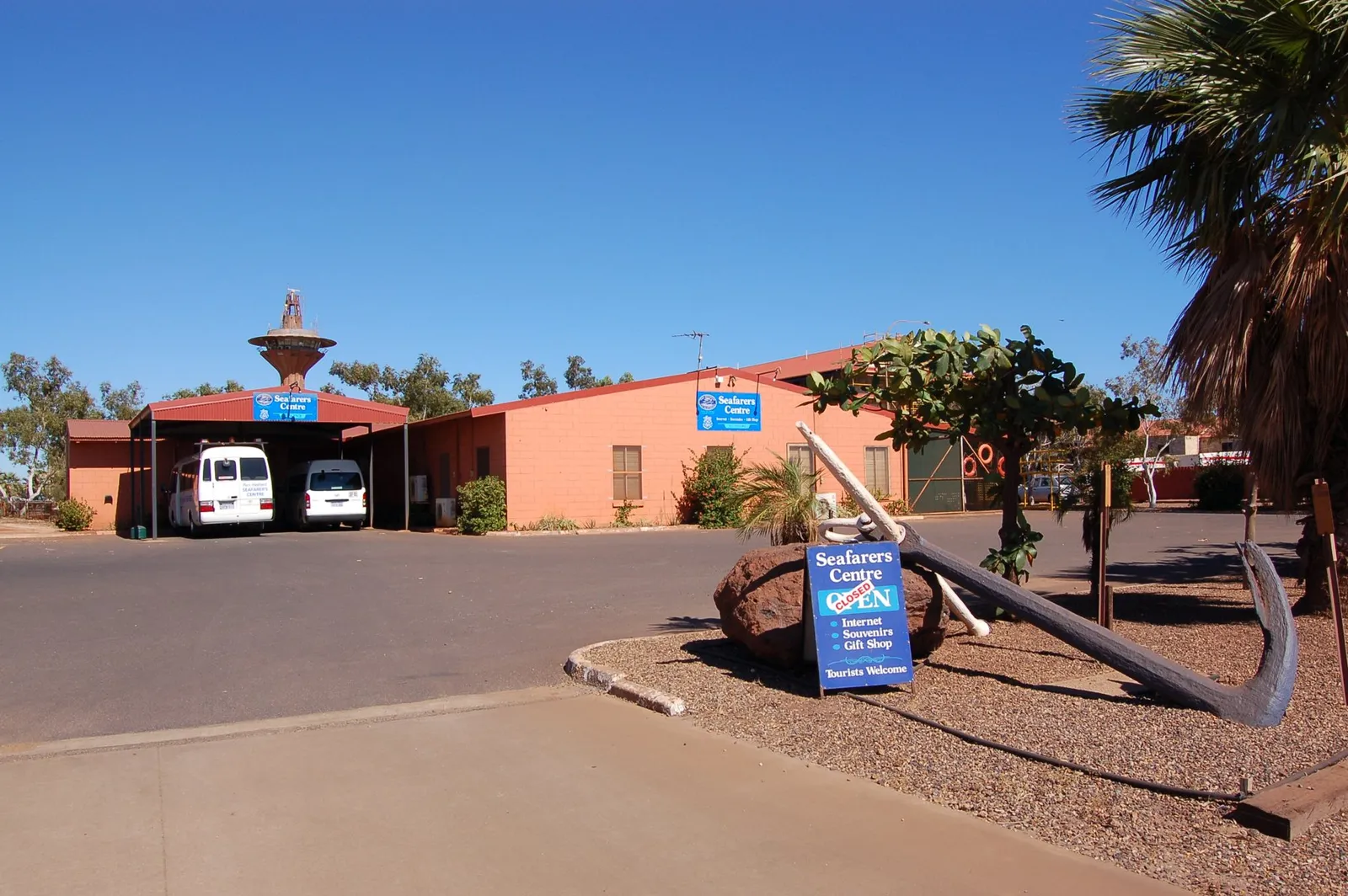A low pink-brick building with blue Seafarers Centre signs, two white minibuses under a carport and a large old anchor beside the entrance, under a clear blue sky.