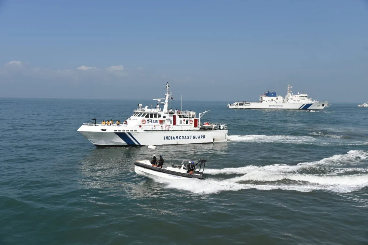 A white Indian Coast Guard patrol boat and a small inflatable boat moving across calm sea, with a larger coast guard ship in the background.