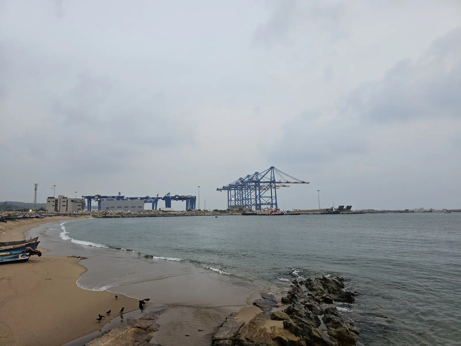 A sandy beach with fishing boats in the foreground and large blue container cranes at a seaport across the water under a grey sky.