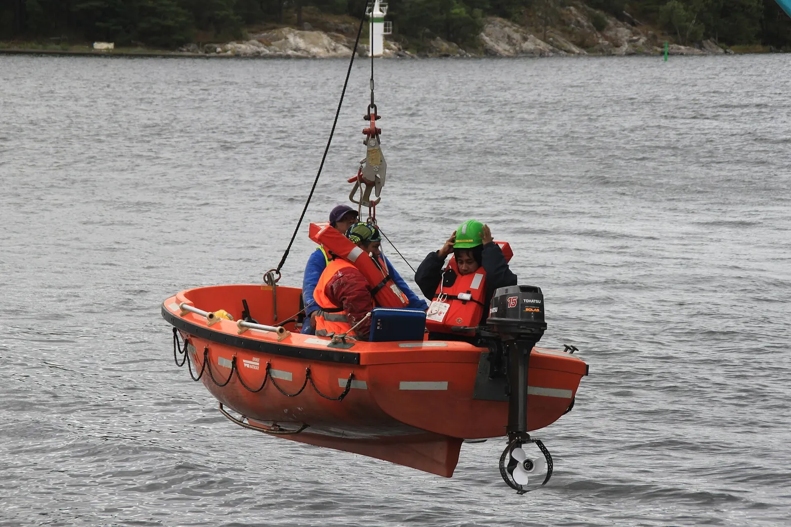 An orange rescue boat with three crew in lifejackets and helmets hanging from a single hoist wire just above the water.