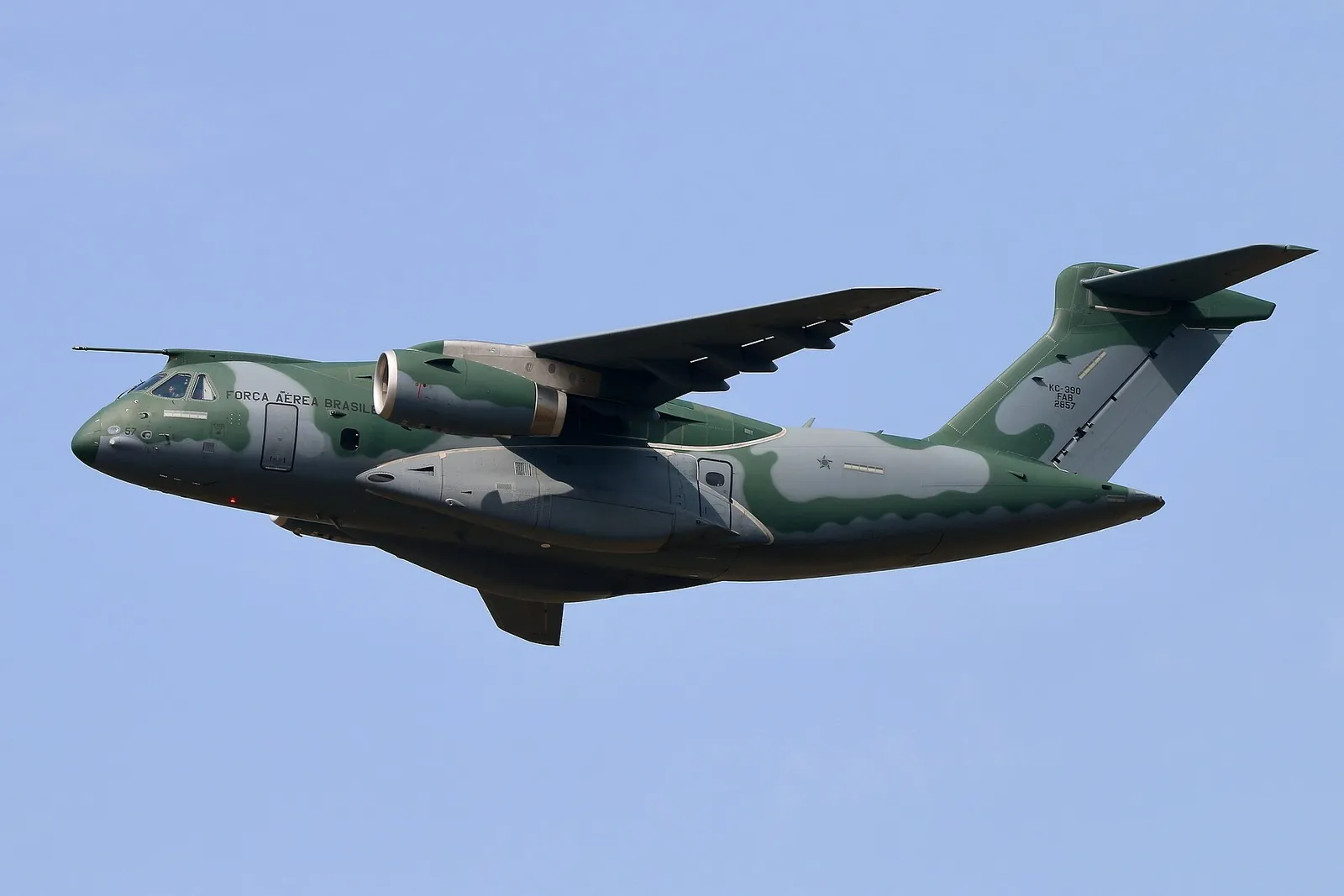 A grey military transport aircraft with high wings and two jet engines flying against a pale sky.