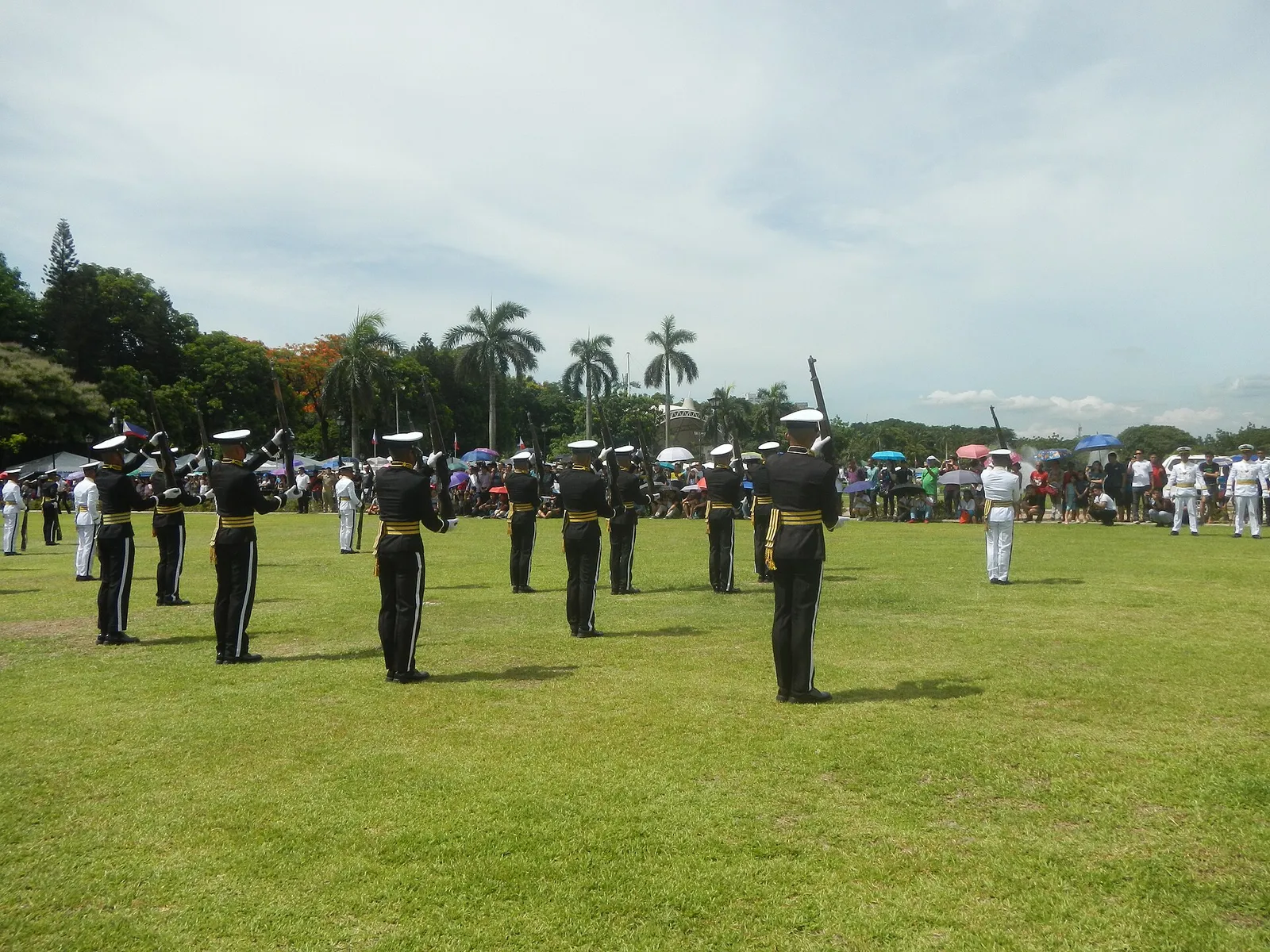 Rows of cadets in black and gold uniforms with rifles on a grass field, with palm trees and spectators behind them.