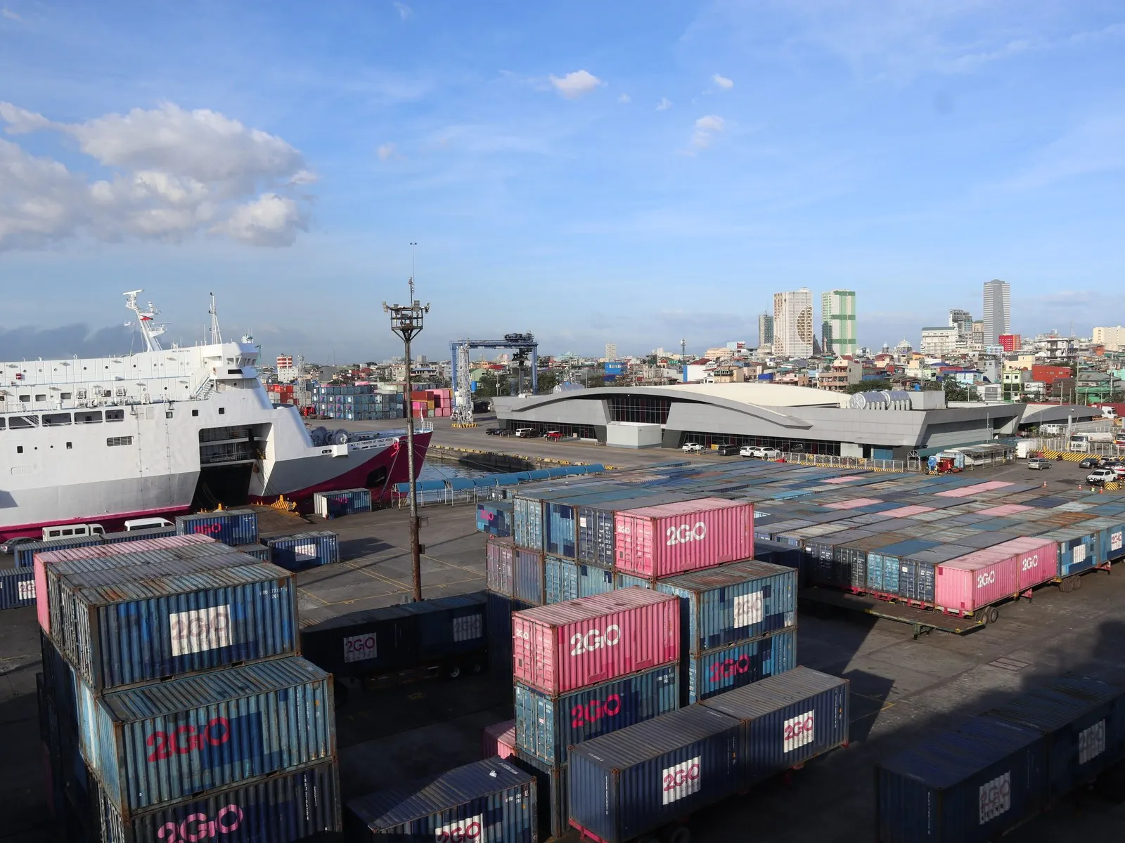 A large white ferry at a pier on the left. Many blue and pink containers stand in the port yard, with city buildings behind.