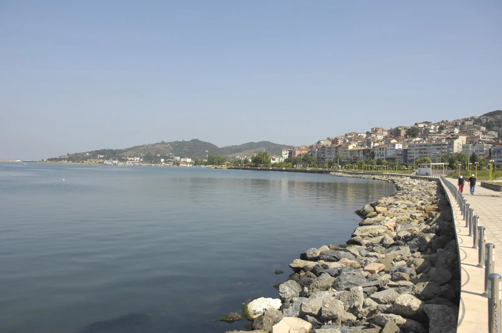 A calm bay with a stone breakwater and promenade in the foreground and a hillside town of white buildings in the distance.