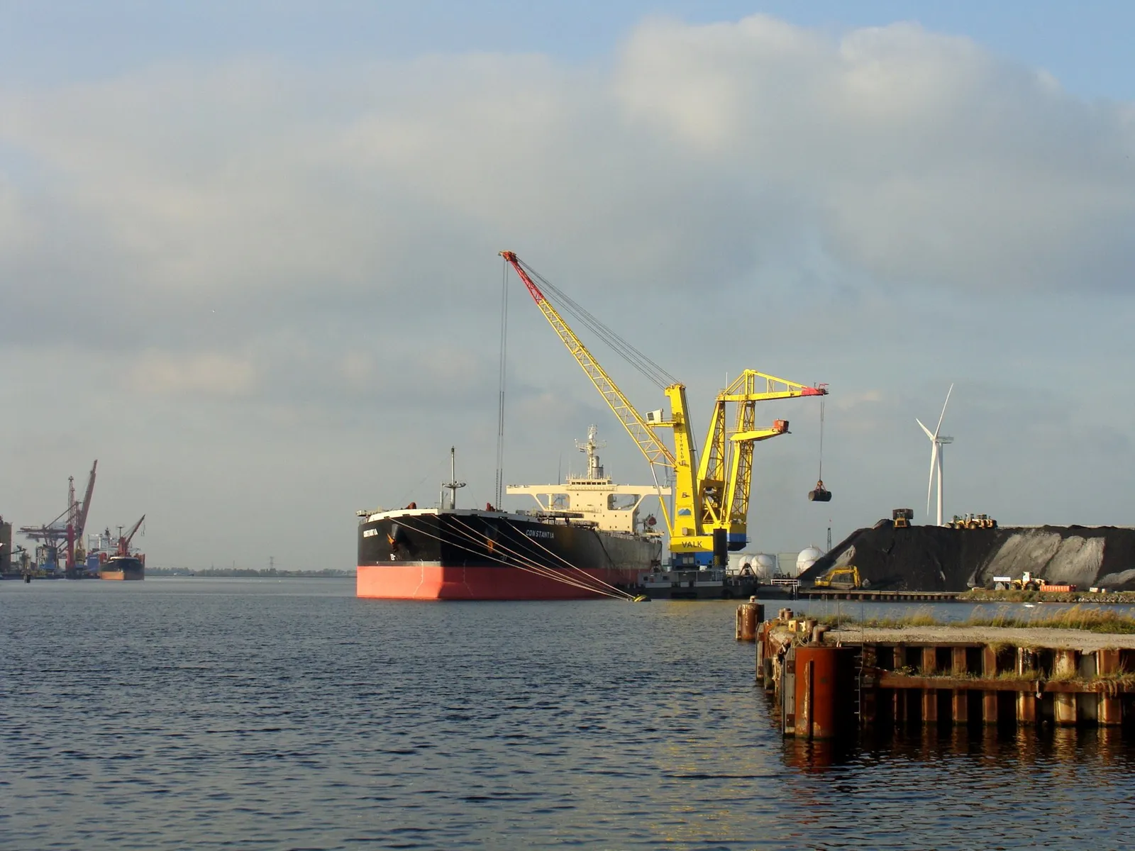 A large bulk carrier with a black hull alongside a yellow grab crane at a coal terminal, with a wind turbine and coal heaps on the right.