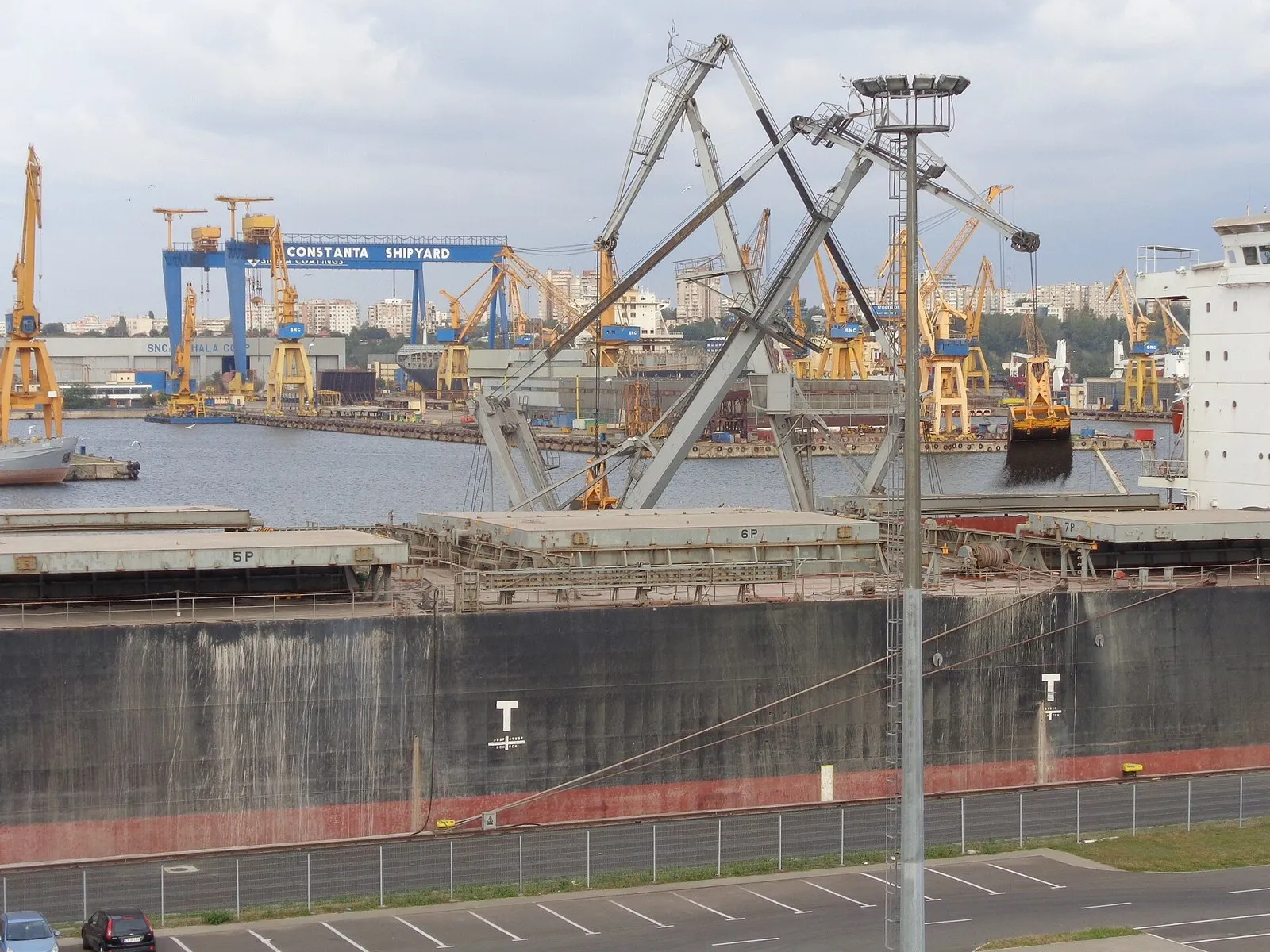 A large dark-hulled bulk carrier alongside a quay, with cranes loading coal into its holds and a shipyard gantry in the background.