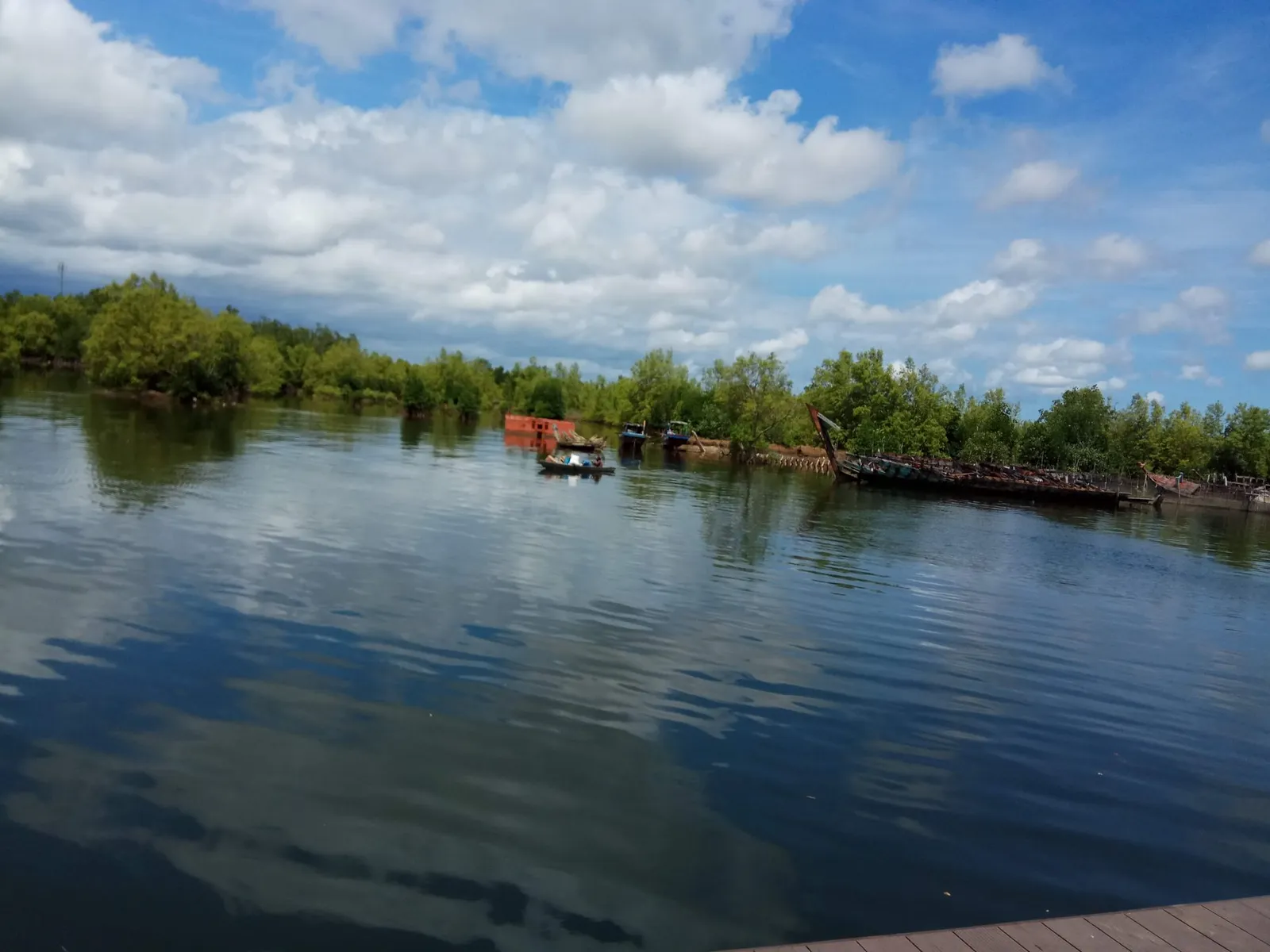 Calm water reflecting clouds, with mangrove trees along the shore, a few small boats and the rusting remains of old vessels.