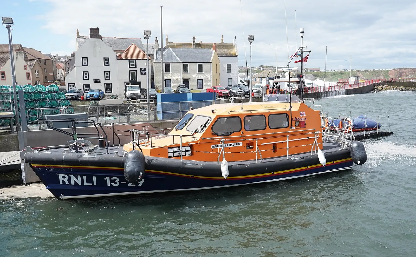 An orange and dark blue RNLI lifeboat with the number 13-29 tied up at a harbour wall, with houses behind it.