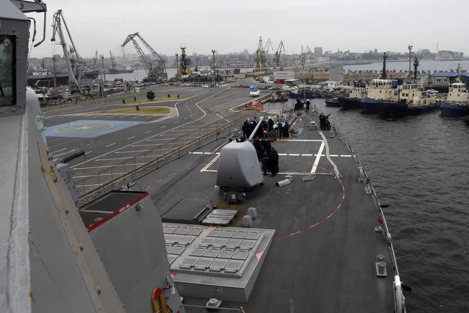View from a warship deck over a busy port with cranes, tugs at a quay and a city in the background under a grey sky.