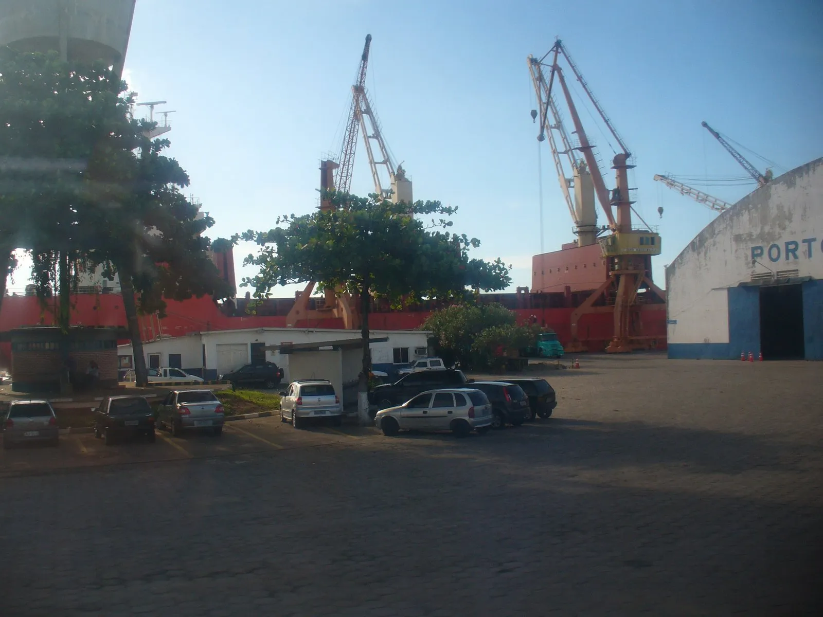 Port cranes stand next to a red ship behind a car park and a white warehouse with the word Porto.