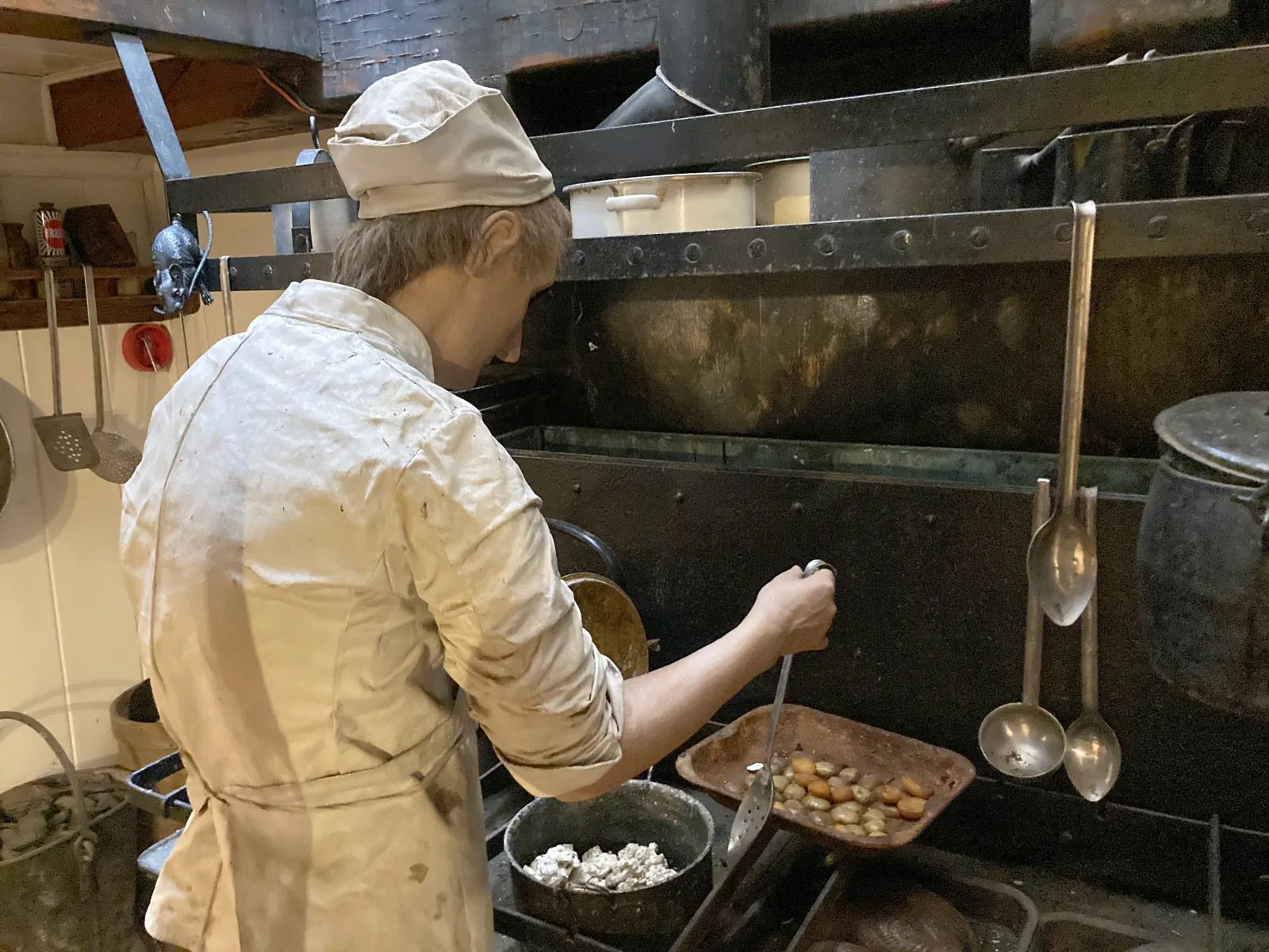 A figure of a cook in a white jacket and cap stirs food on an old iron stove in a ship's kitchen.