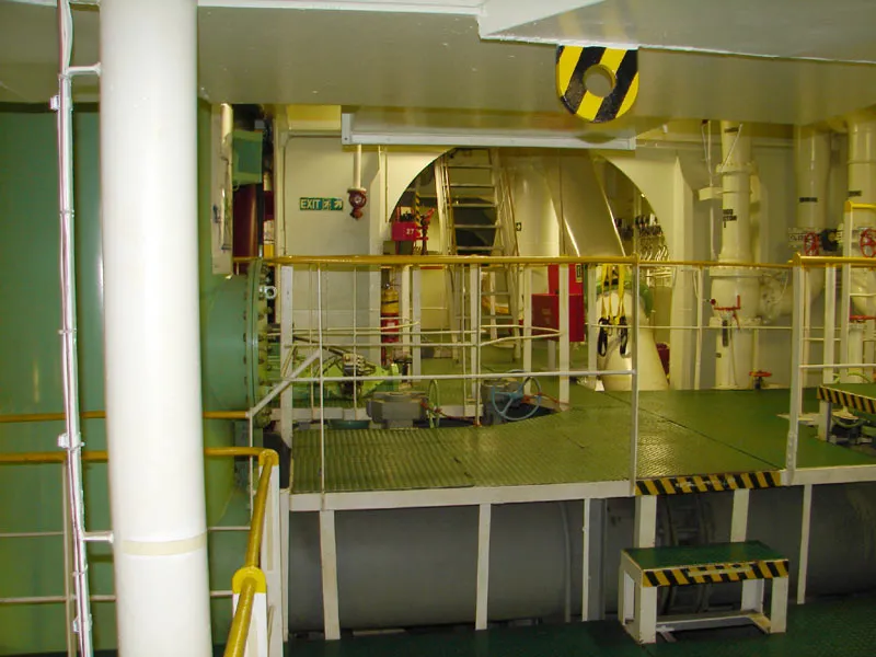 A tidy pump room on a tanker with green floor gratings, yellow handrails, white pipes and a steep ladder leading up.