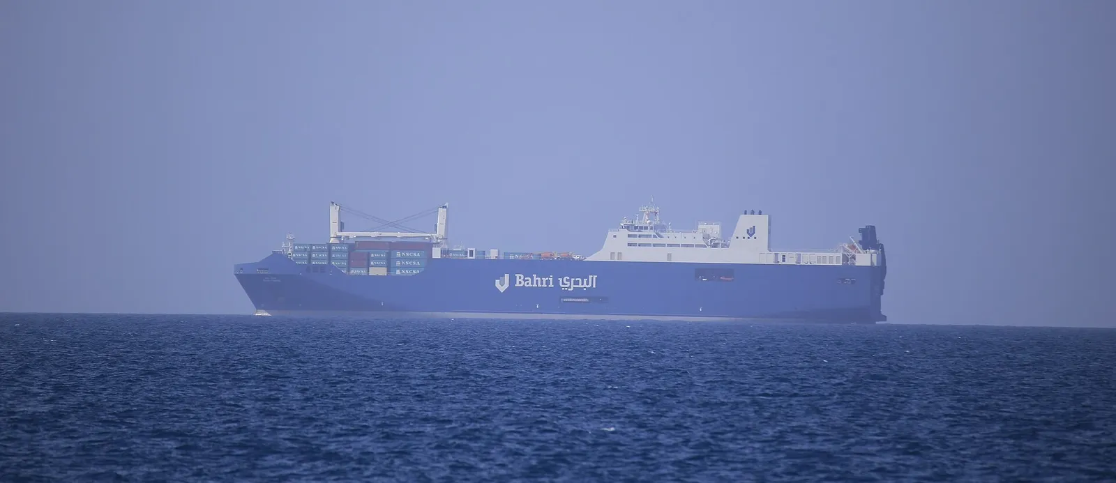 A large blue ro-ro cargo ship with the Bahri name on its side, sailing on a calm blue sea in haze.