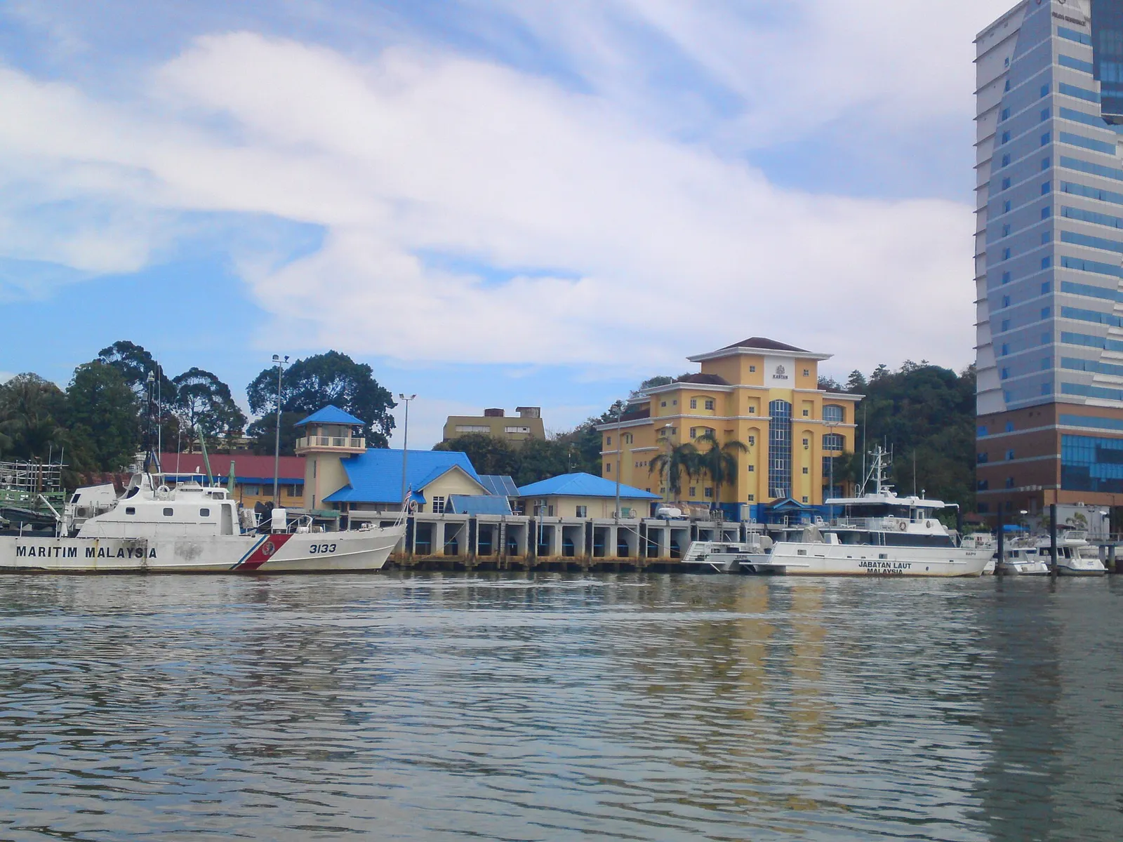 A white patrol ship with Maritim Malaysia written on its side is moored at a jetty in front of yellow and blue buildings.