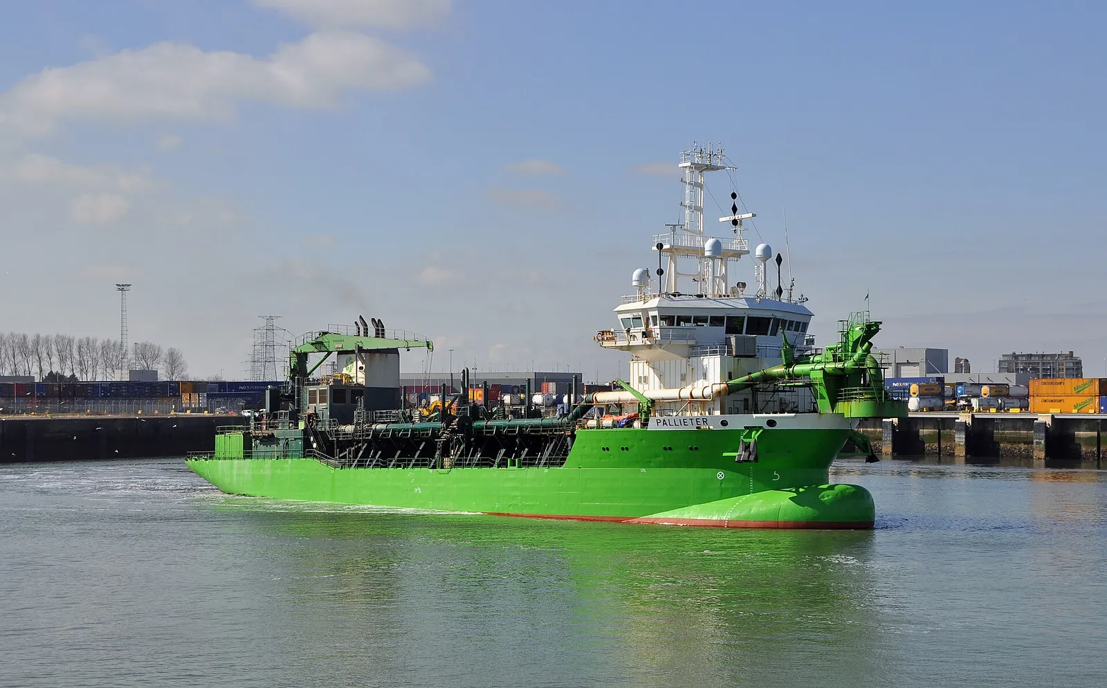 A green dredger with a white superstructure and large pipes on deck sails in a harbour.