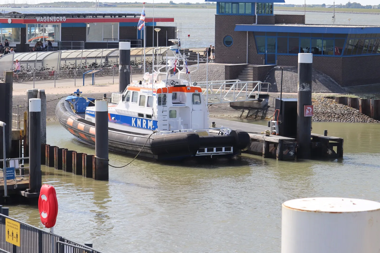 An orange, white and blue KNRM rescue boat moored at a pontoon in a small harbour, with ferry terminal buildings behind it.
