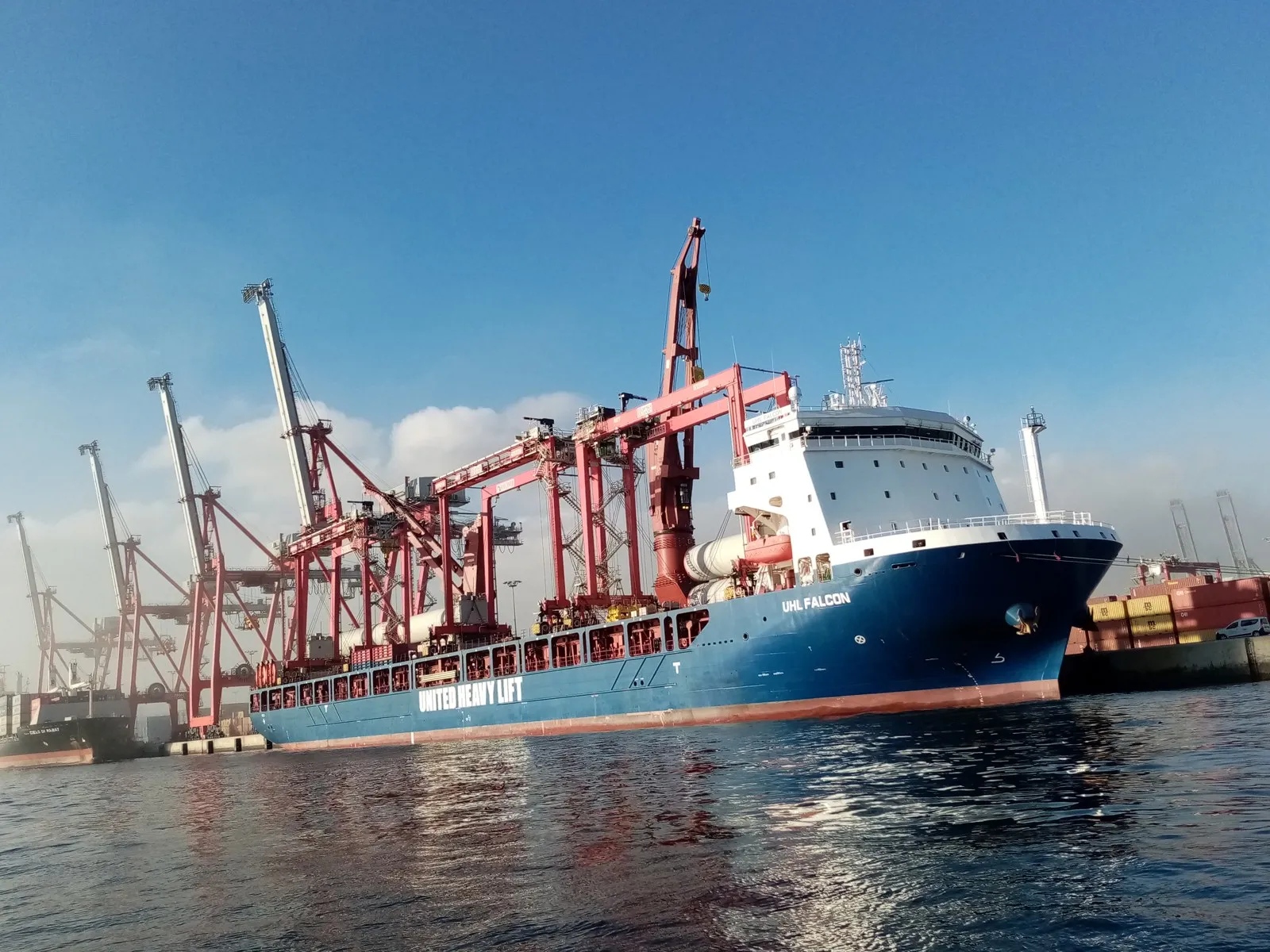 A blue and white cargo ship with large red cranes on deck lies at a quay. The words United Heavy Lift are painted on the hull.