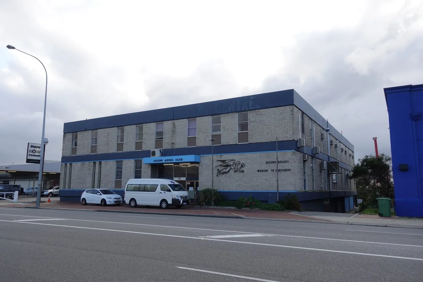 A two-storey brick building with a blue sign reading Flying Angel Club and the words Mission to Seafarers on the wall, with a white minibus parked in front.