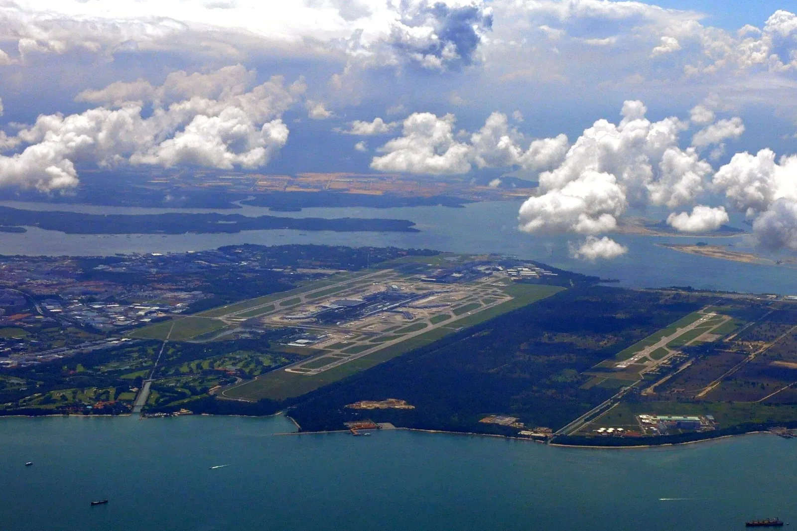 Aerial view of a large airport with long runways next to the sea, with green land and clouds around it.