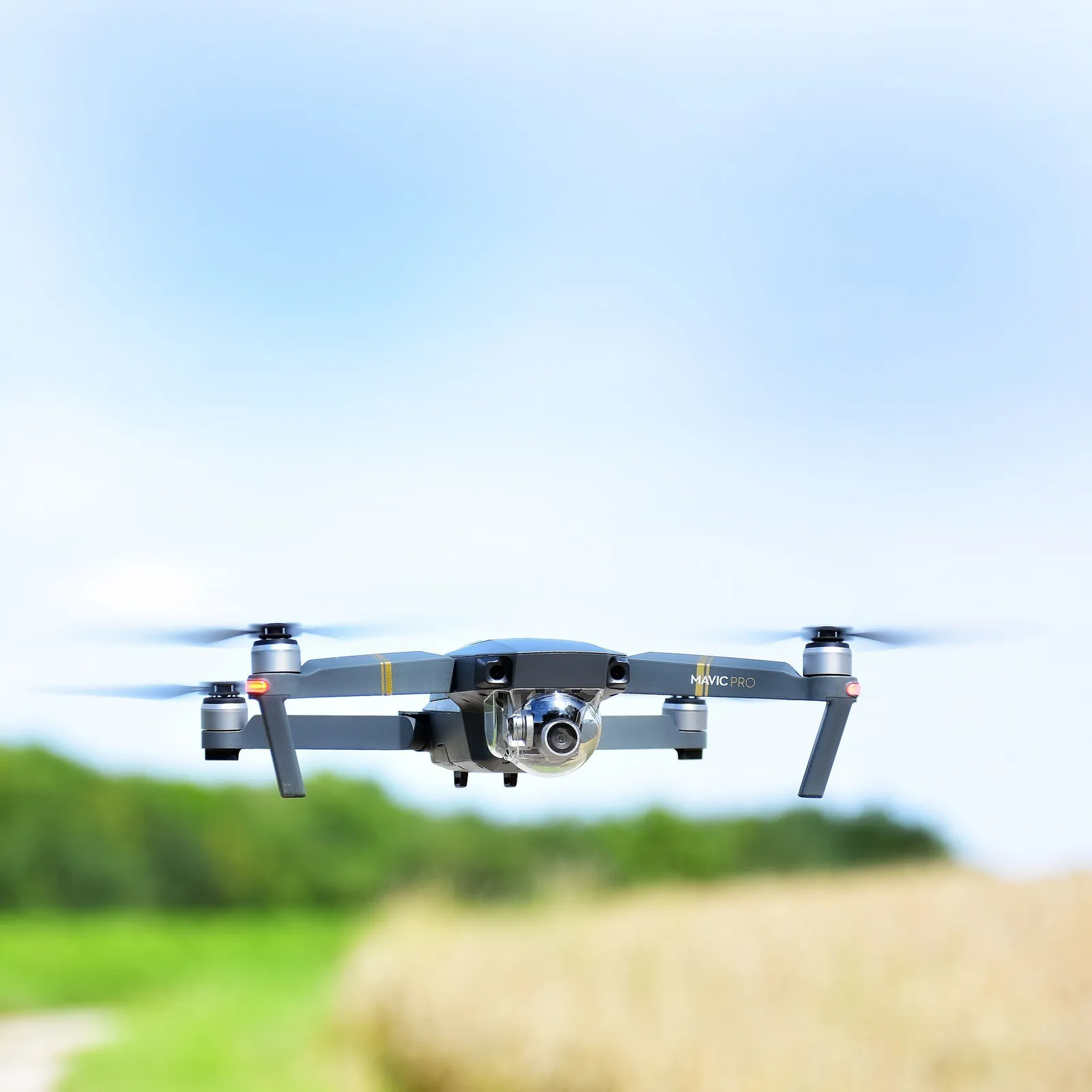 A small grey four-rotor camera drone flying above a field, with trees behind it.