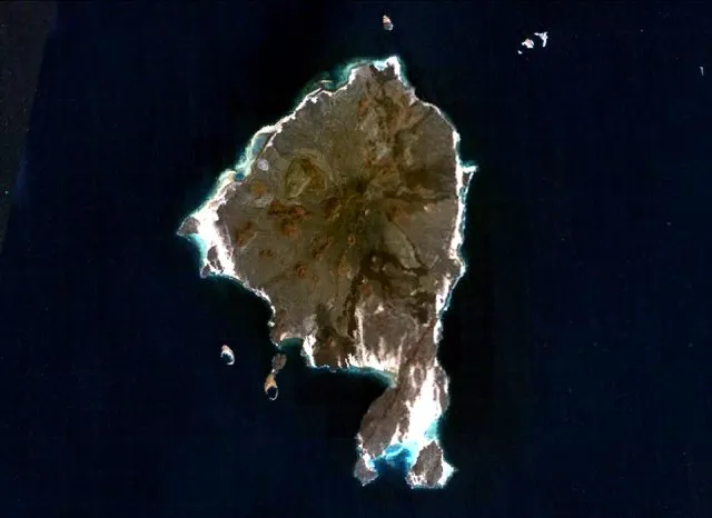 Satellite view of a rocky brown island with white beaches surrounded by dark blue sea.