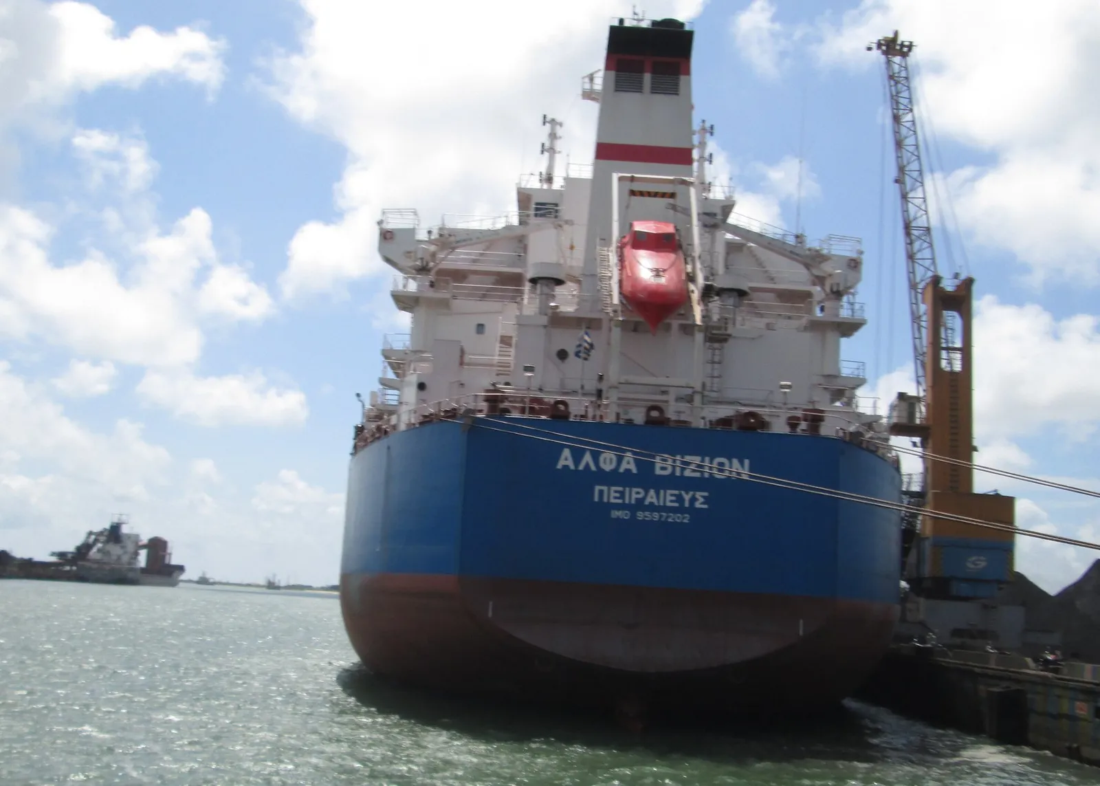 The stern of a large blue bulk carrier with a red lifeboat, moored at a quay next to a tall crane.
