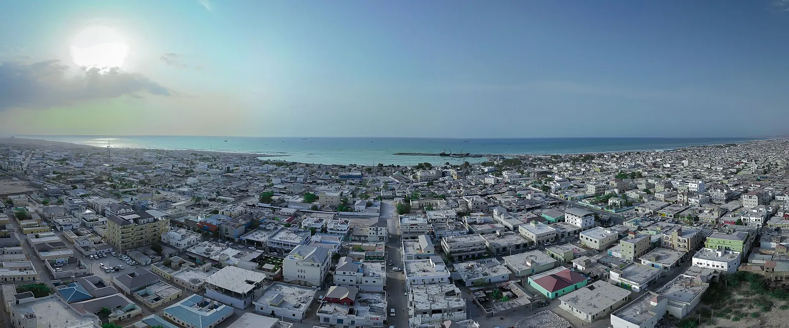 An aerial view of a large low-rise city of white buildings stretching to the sea under a hazy sky.