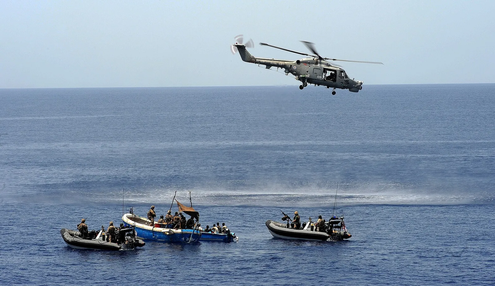 Two grey military boats beside a blue wooden skiff at sea, with a helicopter flying low above them.