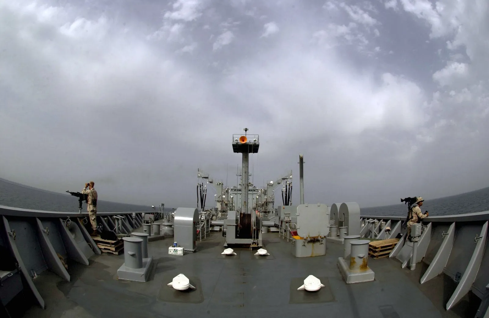 Two security personnel keep watch from the bow of a grey naval supply ship under a cloudy sky.