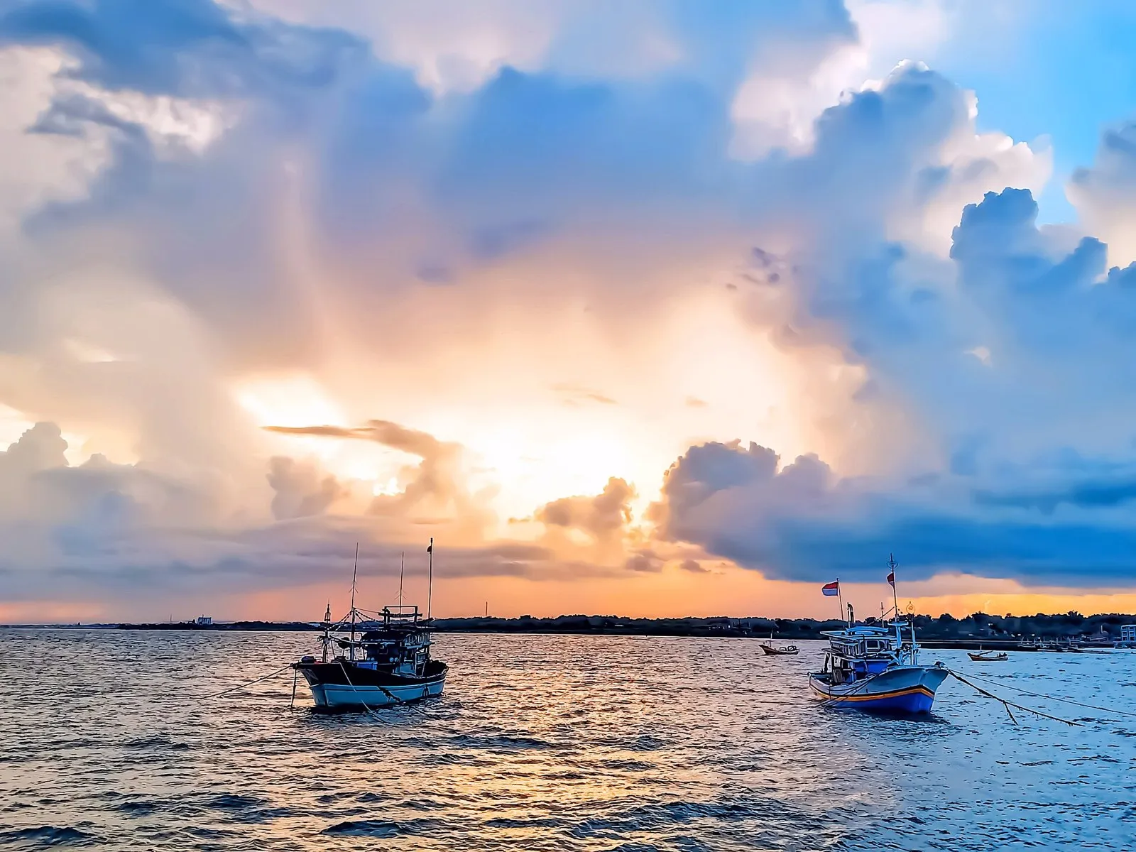 Two small fishing boats at anchor on calm water under a sky of orange and blue clouds at sunset.