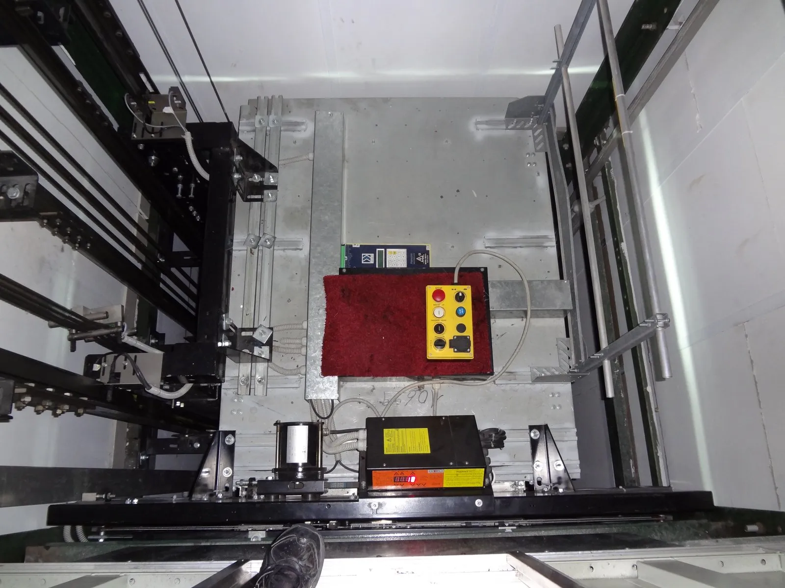 Top of a lift car seen from above, with a yellow control box with a red stop button, ropes and guide rails.