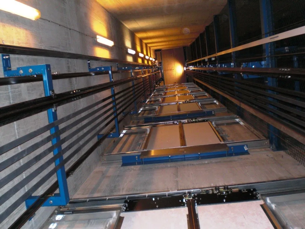 View up the inside of a lift shaft with guide rails, wall lights and a row of closed landing doors.