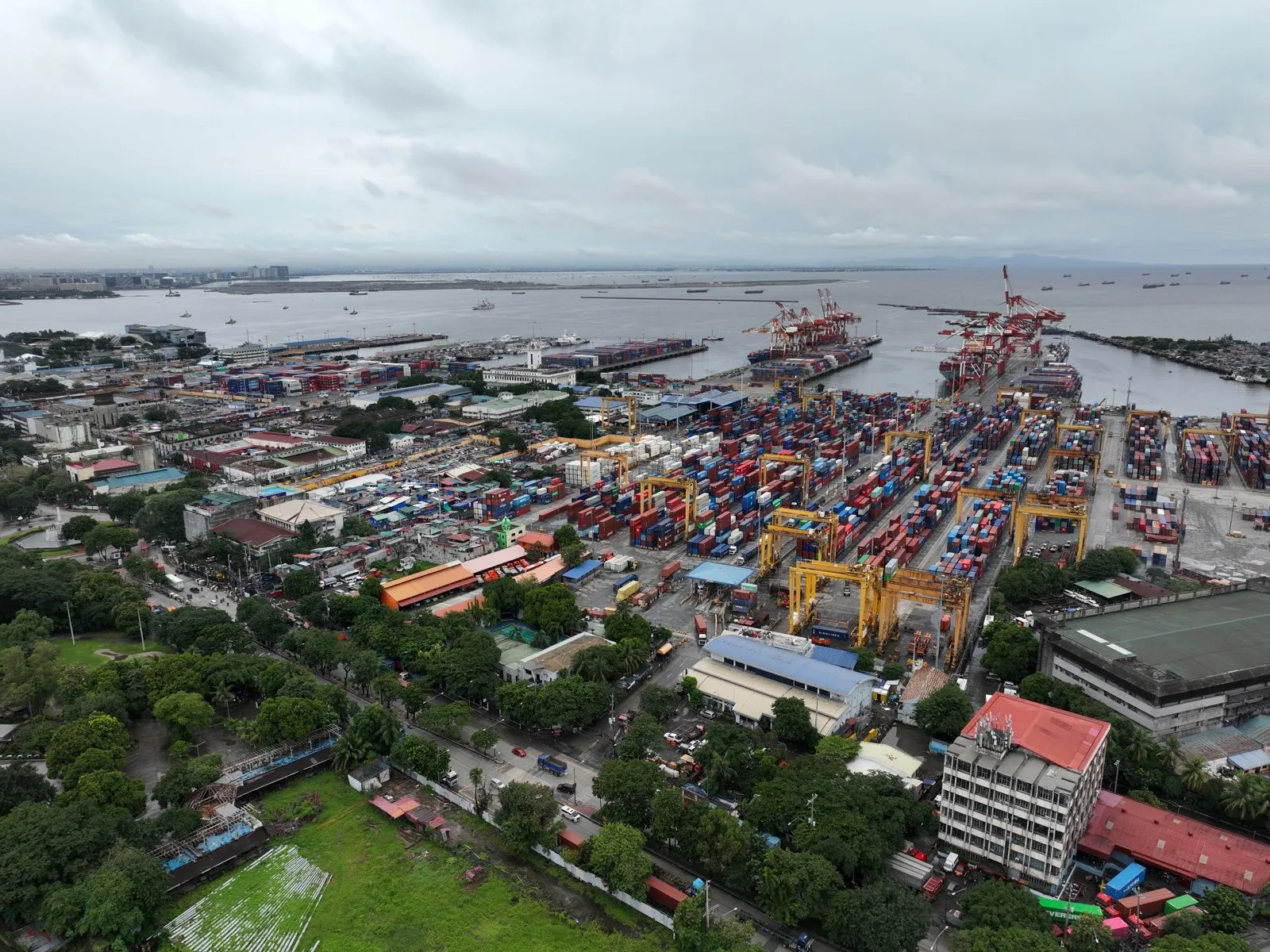 Aerial view of a busy container port with cranes and stacks of containers, the city on the left and Manila Bay behind.