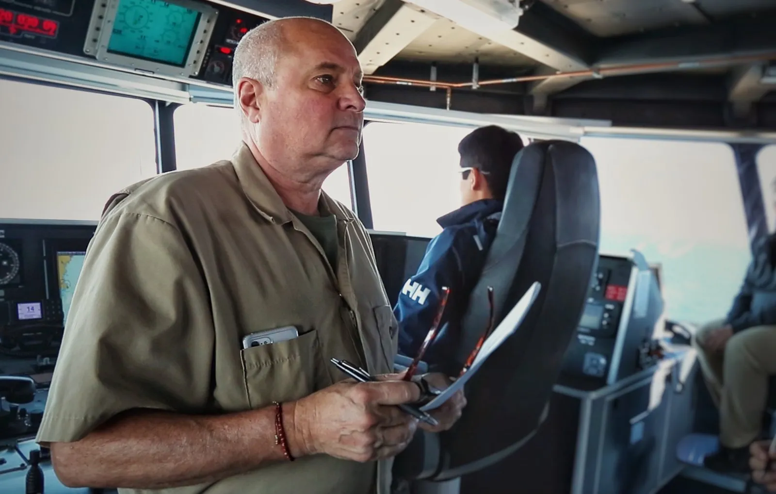 An officer in a khaki shirt stands on a ship's bridge holding papers, with a crew member in a chair behind him.